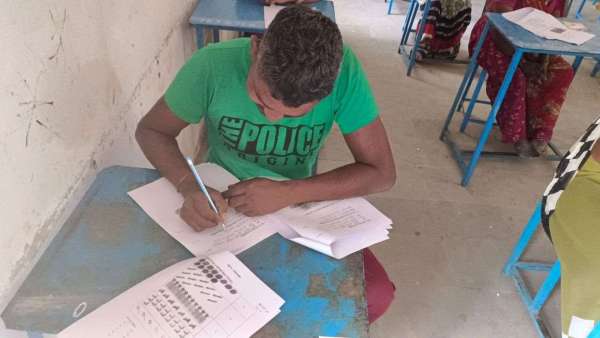 निरक्षर की मूल्यांकन पुस्तिका में उत्तर लिखता कार्मिक।