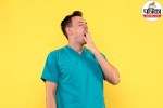 Man in medical scrubs yawning with hand over mouth against yellow background