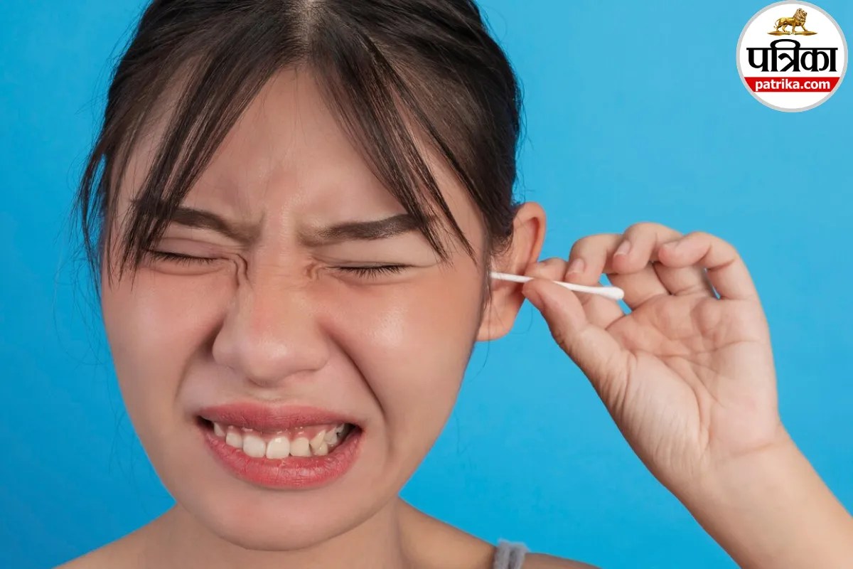 A young woman wincing in pain while inserting a cotton swab inside her ear against a plain blue background.