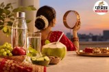 करवा चौथ के लिए सजी महिला के सामने पानी की बोतल, गिलास, नारियल पानी, अंगूर, सेब और खीरे रखे हैं।